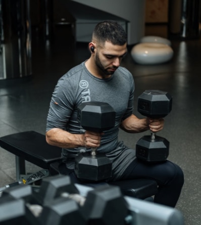 Photo of a young man, with a beard, holding two large dumbbells in a gym.